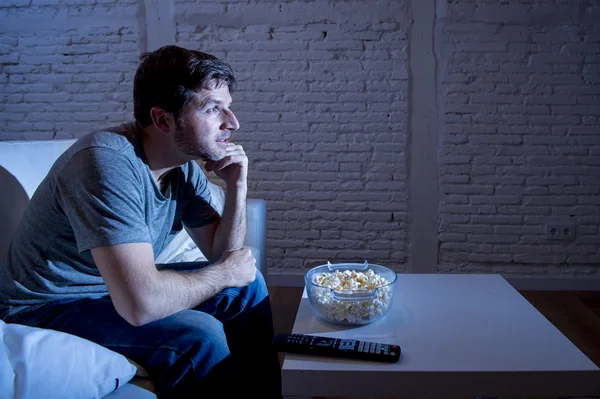 Young happy television addict man sitting on home sofa watching TV and eating popcorn — Stock fotografie