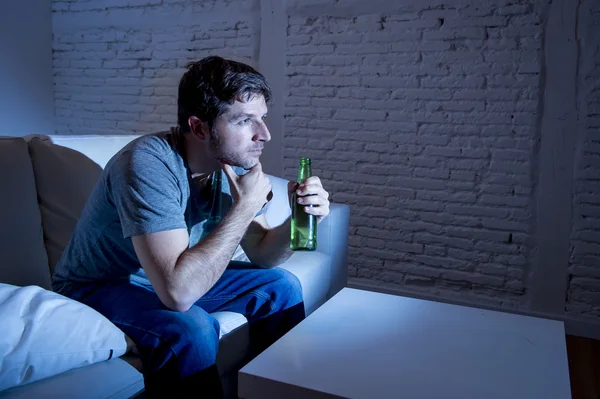 Молодой телевизионный наркоман сидит на домашнем диване и смотрит телевизор и пьет бутылку пива — стоковое фото