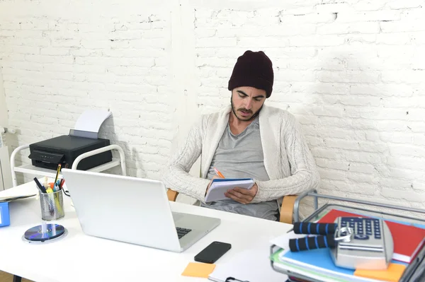 trendy businessman in cool hipster beanie writing on pad working in at modern home office with computer