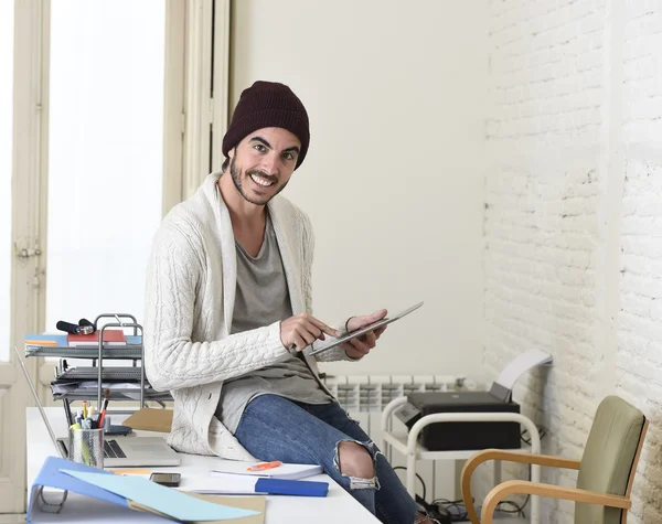 Young trendy businessman in beanie and cool hipster informal look sitting on home office desk using digital tablet pad happy — Stockfoto