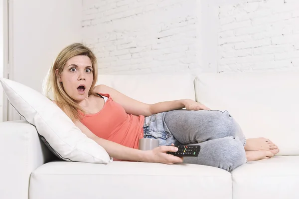 Молода жінка на дивані тримає телевізійний пульт дистанційного керування дивитися телевізор здивована і шокована — стокове фото