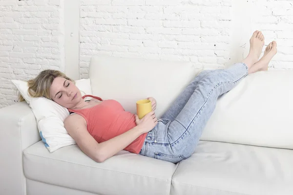 Jovem atraente mulher cabelo loiro segurando xícara de café deitado no sofá sofá em casa sala de estar dormindo relaxado — Fotografia de Stock