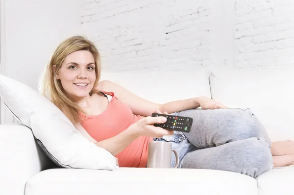 Wanita muda di sofa memegang remote control televisi menonton TV tersenyum bahagia — Stok Foto