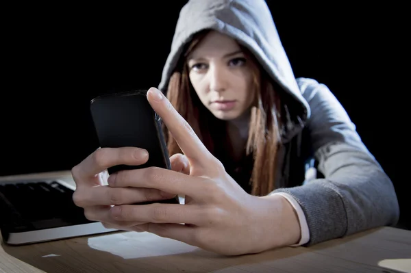 Adolescente hacker chica en la capucha usando el teléfono móvil en internet experto en delitos cibernéticos o cibercrimen —  Fotos de Stock