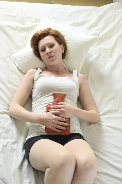Молодая женщина страдает спазмами желудка на животе держа бутылку с горячей водой против живота — стоковое фото