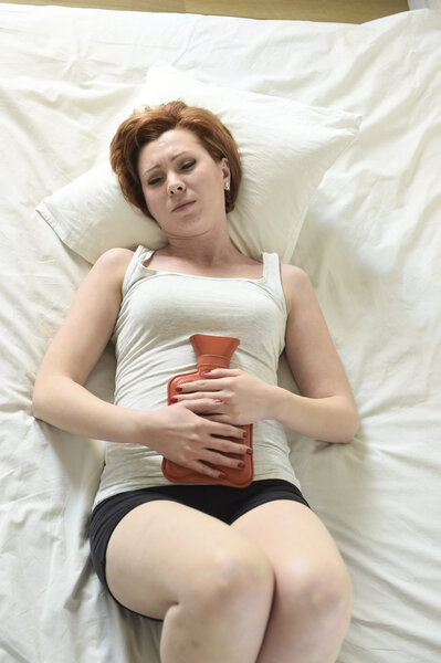 young woman suffering stomach cramps on belly holding hot water bottle against tummy