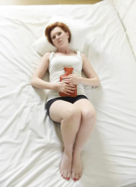 Young woman suffering stomach cramps on belly holding hot water bottle against tummy — ストック写真