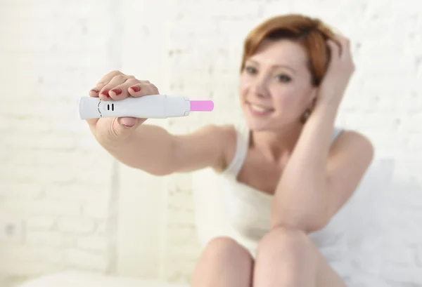 Young attractive pregnant woman holding happy positive result pregnancy test sitting on bed — Stockfoto