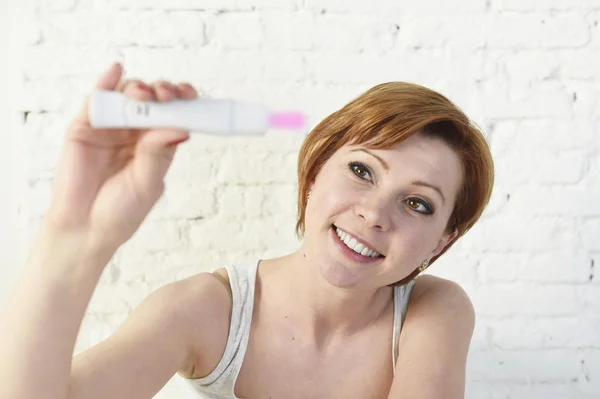 Young attractive pregnant woman holding happy positive result pregnancy test sitting on bed — Stock fotografie