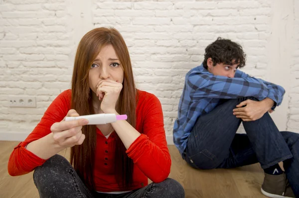 young sad couple scared in shock and surprise reading pink positive pregnancy test overwhelmed in panic