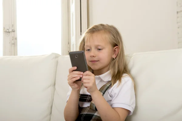 Niña con cabello rubio sentada en el sofá utilizando la aplicación de Internet en el teléfono móvil — Foto de Stock