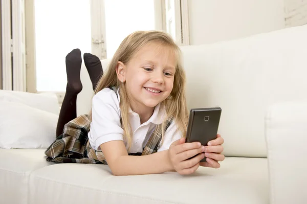 Female child with blond hair in school uniform lying on home sofa couch using internet app on mobile phone — Stock Photo, Image