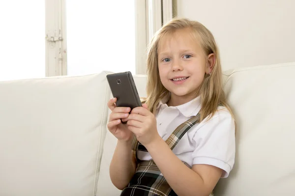 Criança do sexo feminino com cabelo loiro sentado no sofá usando o aplicativo de internet no telefone móvel — Fotografia de Stock
