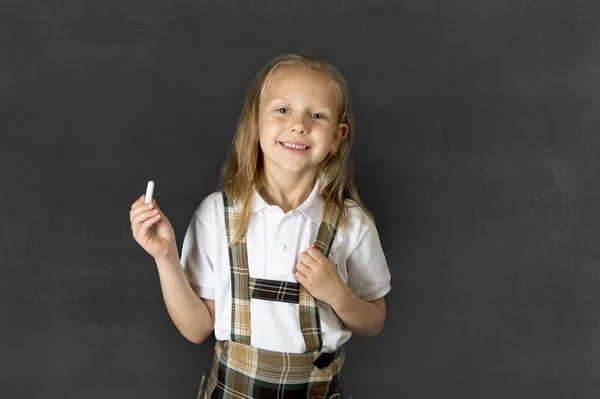 Sweet junior schoolgirl with blonde hair standing and smiling happy isolated in blackboard background — Stock Photo, Image