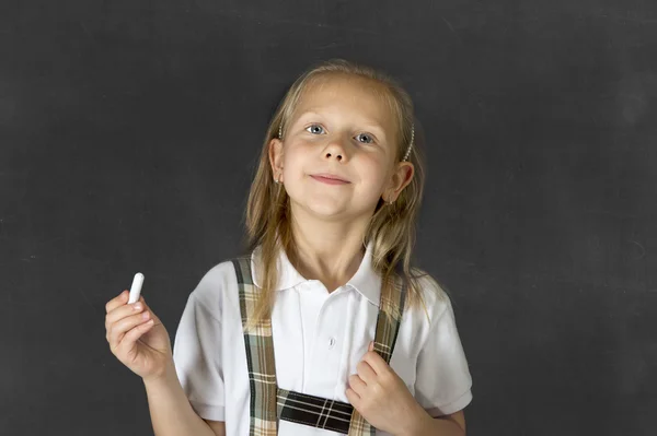 Söta junior skolflicka med blont hår står och ler lyckligt isolerade i blackboard bakgrund — Stockfoto
