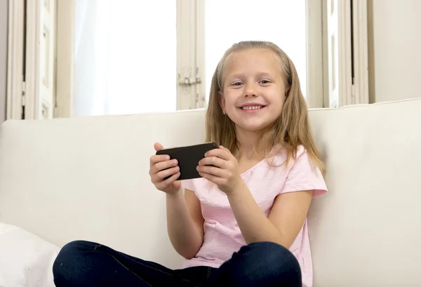 Happy blond little girl on home sofa using internet app on mobile phone — Stock Photo, Image