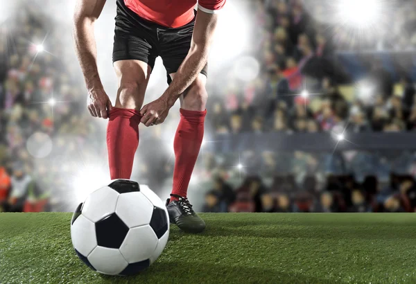 Футболист с мячом в черных ботинках регулирует свой красный носок стоя на стадионе поле — стоковое фото