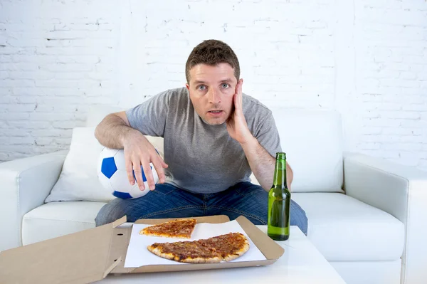Young man alone holding ball and beer bottle watching football game on television at home sofa couch — Stock Photo, Image