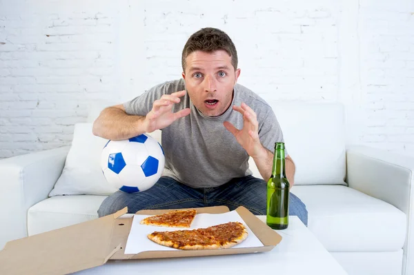 Ung man ensam håller i bollen och öl flaska titta på fotbollsmatch på TV på hem soffa soffa — Stockfoto