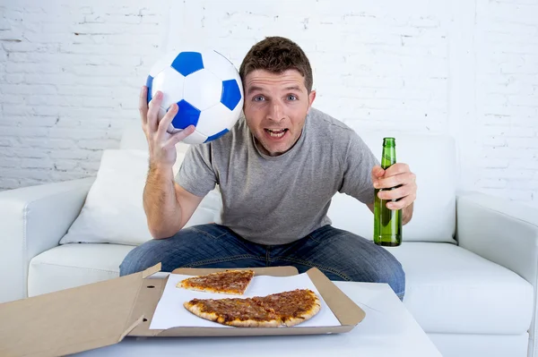 Mladý muž sám drží míč a pivní láhev sledovat fotbalový zápas v televizi na domácí rozkládací gauč — Stock fotografie