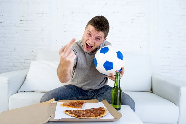 Joven sosteniendo pelota viendo partido de fútbol en la televisión en el sofá de casa con cerveza celebrando loco dando el dedo —  Fotos de Stock