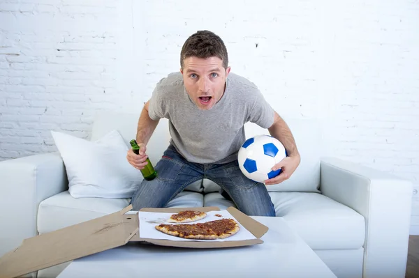 Młody człowiek sam trzymając piłkę i piwo butelka oglądania meczu piłki nożnej w telewizji w domu sofa kanapa — Zdjęcie stockowe