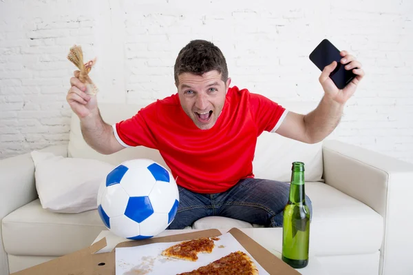 Молодой человек держит в руках мобильный телефон и деньги, наблюдая за игрой в настольный футбол по телевидению — стоковое фото