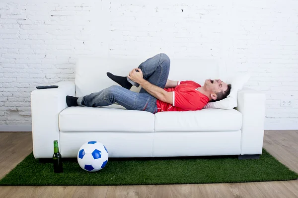 Фанат футбола лежит на диване с мячом на зеленом травяном ковре, подражая футбольному стадиону, насмехаясь над игроком, страдающим от боли в лодыжке. — стоковое фото