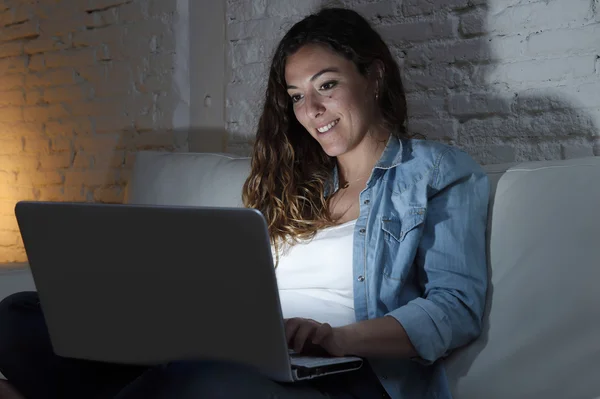 Ελκυστική γυναίκα χαλαρή στο σπίτι κάθεται ευτυχισμένος στον καναπέ χρησιμοποιώντας φορητό υπολογιστή σκοτεινό βράδυ — Φωτογραφία Αρχείου