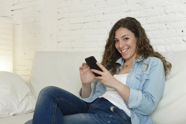 Jovem mulher espanhola atraente usando aplicativo de telefone móvel ou mensagens de texto no sofá em casa — Fotografia de Stock