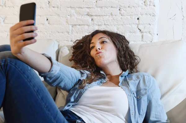 Young attractive hispanic woman lying on home couch taking selfie photo with mobile phone having fun — Zdjęcie stockowe