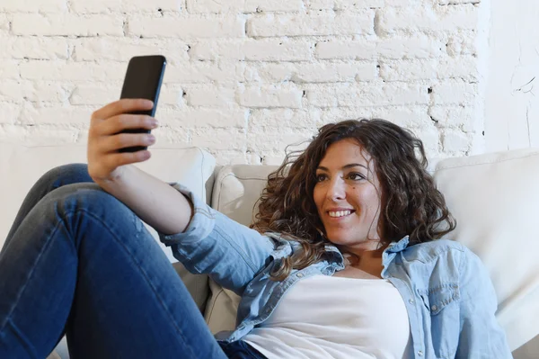 Joven atractiva mujer hispana tumbada en el sofá de casa tomando foto selfie con el teléfono móvil divertirse —  Fotos de Stock