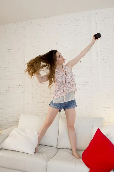 Mutlu çekici kız ev kanepede oynarken cep telefonu ile selfie portre alarak eğlenmek — Stok fotoğraf