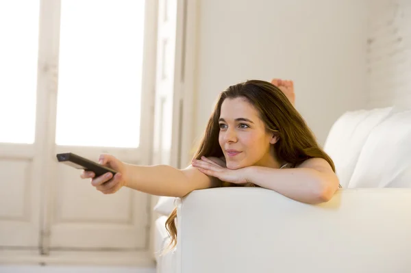 Junge schöne Frau auf dem Sofa liegend mit Fernbedienung vor dem Fernseher glücklich und entspannt — Stockfoto