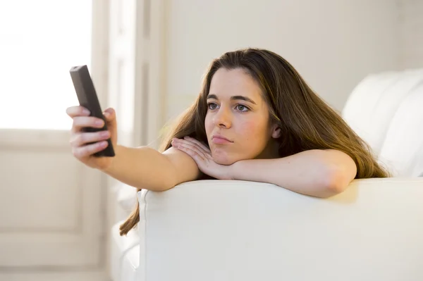 Young beautiful woman lying on couch holding remote control zapping while watching television — Stock Fotó