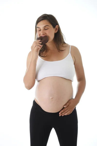 Joven atractiva embarazada sosteniendo su gran vientre con la mano y comiendo donut de chocolate — Foto de Stock