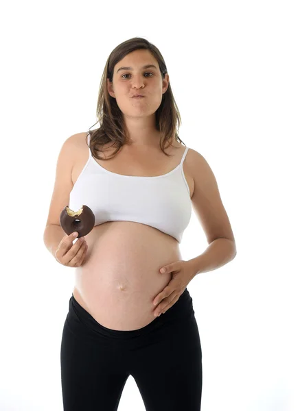 Joven atractiva embarazada sosteniendo su gran vientre con la mano y comiendo donut de chocolate — Foto de Stock