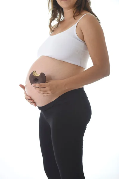 Молодая привлекательная беременная женщина держит большой живот рукой и ест шоколадный пончик — стоковое фото