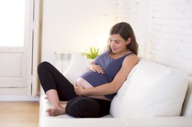 young beautiful happy 8 or 9 months pregnant woman at home living room couch holding big belly