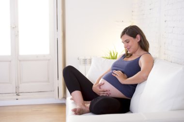 young beautiful happy 8 or 9 months pregnant woman at home living room couch holding big belly