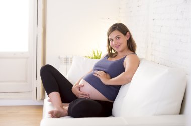 young beautiful happy 8 or 9 months pregnant woman at home living room couch holding big belly