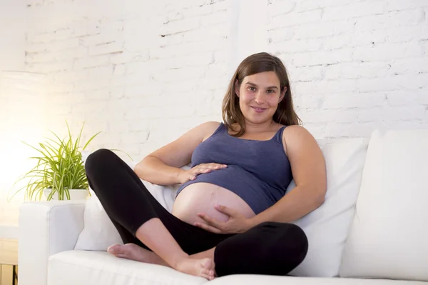 Young beautiful happy 8 or 9 months pregnant woman at home living room couch holding big belly — 图库照片