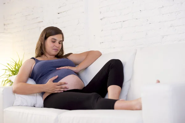 Young beautiful happy 8 or 9 months pregnant woman at home living room couch holding big belly — 图库照片