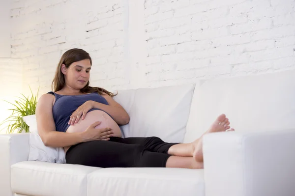 Young beautiful happy 8 or 9 months pregnant woman at home living room couch holding big belly — Stok fotoğraf