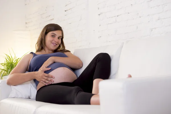 Young beautiful happy 8 or 9 months pregnant woman at home living room couch holding big belly — 图库照片