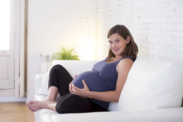 Young beautiful happy 8 or 9 months pregnant woman at home living room couch holding big belly — Stok fotoğraf