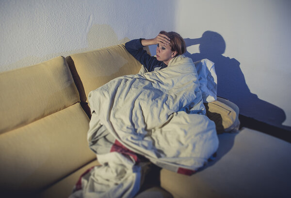 young sick woman sitting on couch wrapped in duvet and blanket feeling miserable