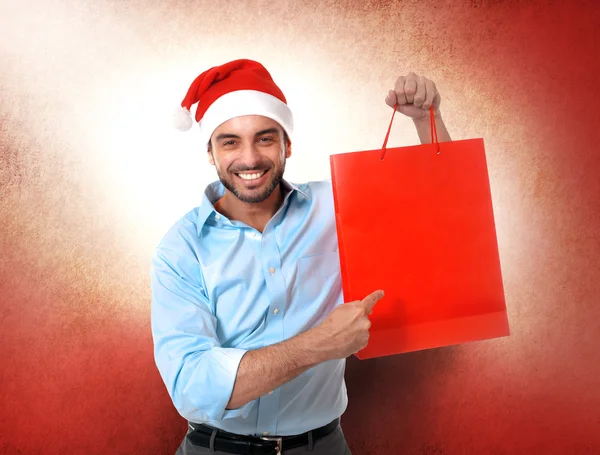 Feliz jovem bonito homem vestindo santa chapéu segurando vermelho compras — Fotografia de Stock