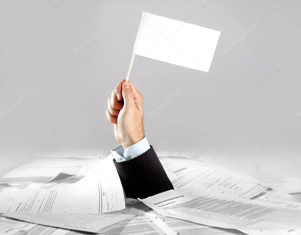 Hand of  businessman emerging from loaded paperwork desk holding white flag