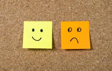 post it notes on corkboard with smiley and sad cartoon face expression in happiness versus depression concept
